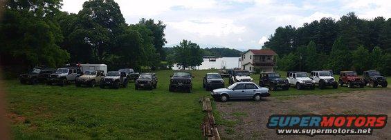 light blue sedan parked in front of several SUVs and Jeeps on a grassy field near a lake