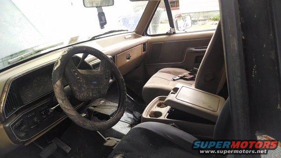 1987 Ford Bronco interior, tan dashboard, dirty steering wheel, open center console with cup holders, worn seats