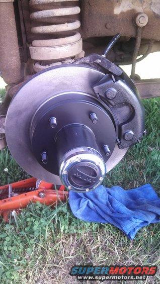 close-up of a vehicle's front brake rotor, caliper, and hub with a "LOCK" hubcap. A jack is visible below