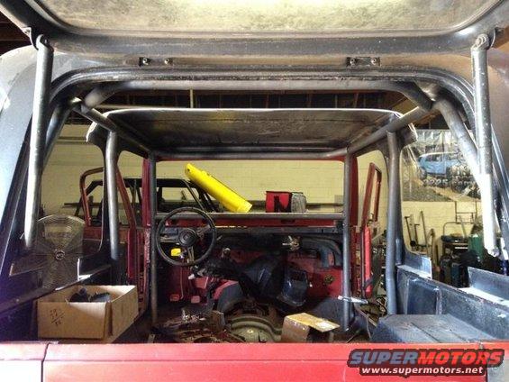 Interior of a red Jeep with a roll cage, dashboard, and steering wheel visible. Another Jeep is in the background
