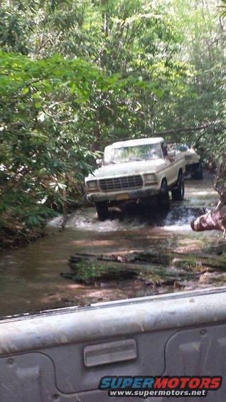 White 1979 Ford F-150 pickup truck driving through a shallow stream with trees on both sides. Another vehicle is visible behind it