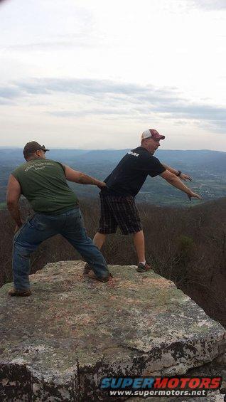 Two men stand on a large rock formation, looking out over a distant valley and hills under a cloudy sky