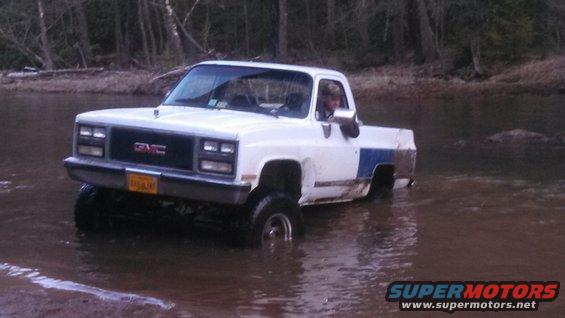 White GMC pickup truck with blue stripe driving in shallow water, driver visible