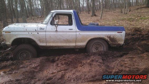 White and blue Ford Bronco stuck deep in mud on a wooded trail