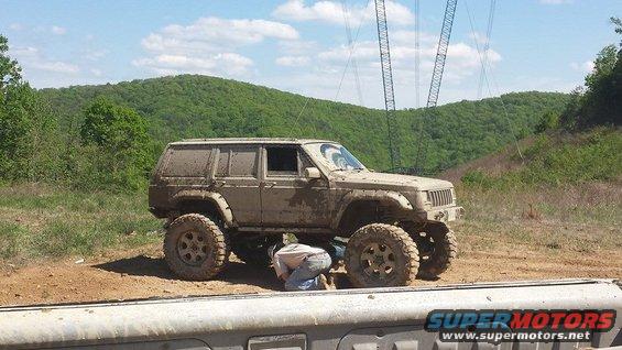 Mud-covered Jeep Cherokee XJ with large tires, being worked on by a person underneath