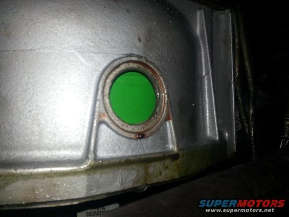 Close-up of a transmission housing with a circular opening showing green fluid. Some oil is visible around the housing