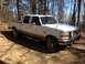 1988 Ford Bronco - This white crew cab pickup truck is parked on a dirt road surrounded by trees.