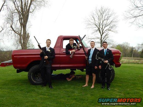 Four guys in suits stand around a red pickup truck, two holding rifles, one holding a bow