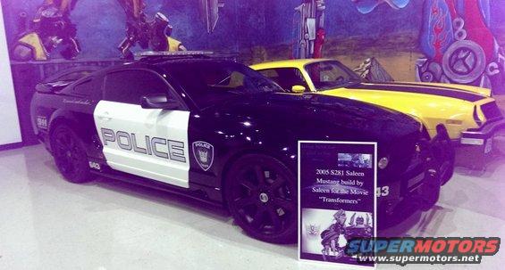 Black and white police car with 'POLICE' on door, parked next to a yellow car with black stripes. Sign in front of police car