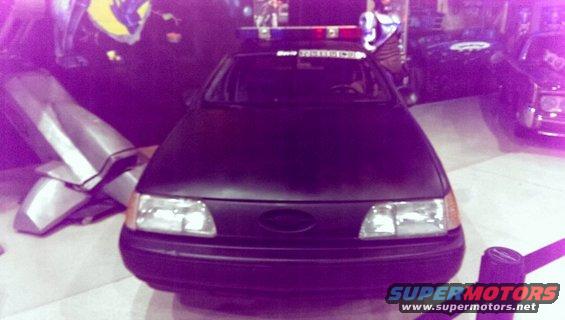 Front view of a dark police car with a light bar on top. Another car is visible to the right
