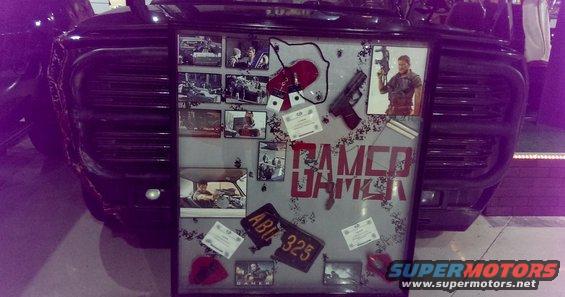Display board with photos, a gun, and 'GAMER' text, mounted on a dark vehicle's grille