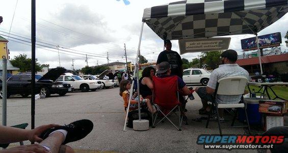 People gathered under a checkered tent at a car show, with several cars parked in the background. A 2000 Mercury Grand Marquis is visible