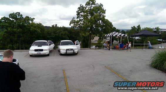 Two white Ford Crown Victoria Police Interceptors parked in a lot, one with hood open. People are gathered under tents in the background
