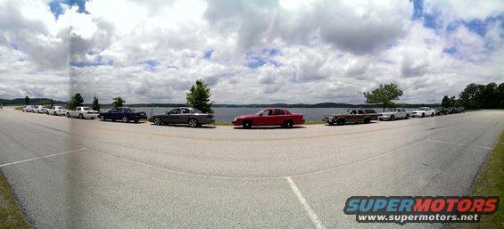 Line of sedans and a station wagon parked on asphalt by a lake under a cloudy sky