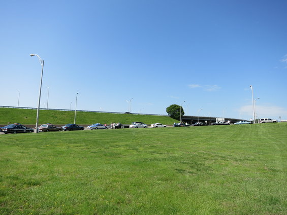 Several cars parked on a grassy slope next to a highway overpass under a clear blue sky