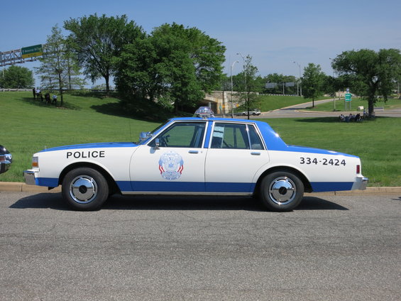 police car with blue and white paint, "POLICE" text on door, and number "334-2424" on rear quarter panel