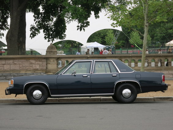 Dark blue sedan parked on the side of the road with a stone wall and trees in the background