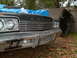 1974 Dodge Monaco - The front of a vintage Dodge sits in a yard, partially covered and with a camper shell leaning nearby.
