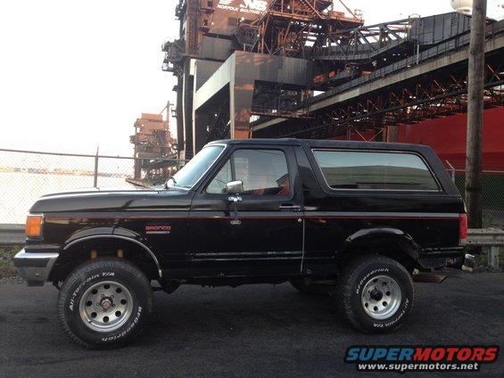 Black Ford Bronco with red pinstripes, side view, parked on asphalt in front of industrial structures