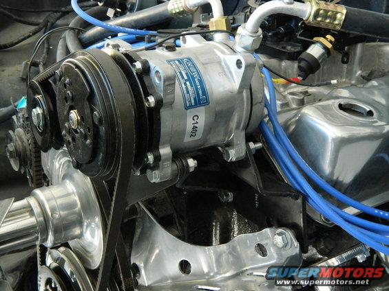 Engine bay showing a silver A/C compressor with blue hoses and a black belt, mounted on a black bracket