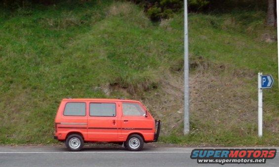 Orange van parked on the side of the road next to a grassy hill and a parking sign
