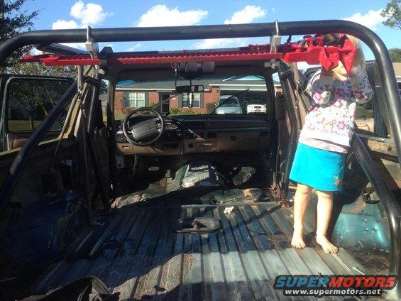 Interior of a modified SUV with a roll cage, stripped rear cargo area, and dashboard visible. A child stands in the rear