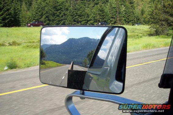 Side mirror reflection shows a mountain road with trees and sky