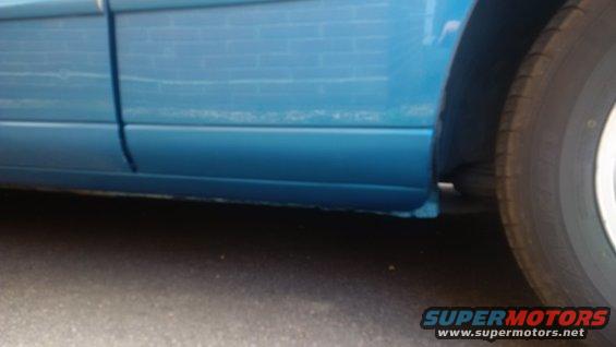 Blue car lower body panel with visible rust repair under the wheel well. A tire is partially visible