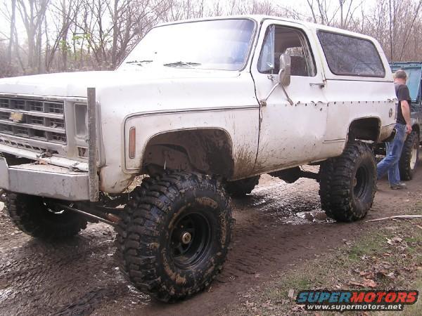 White SUV with lifted suspension and large off-road tires, mud splattered on body. Front grille visible