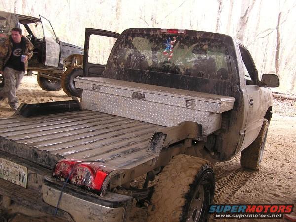 Rear view of a dirty pickup truck with a toolbox in the bed. Another vehicle is visible to the left