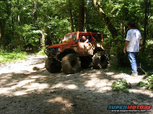 Orange off-road vehicle with large tires on a dirt path in a wooded area. A man stands nearby