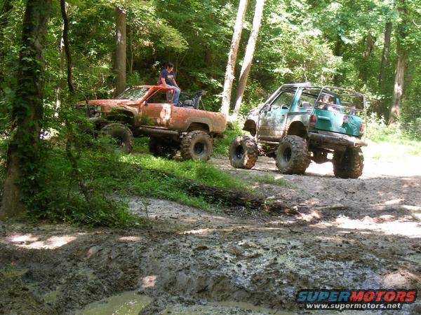 Orange off-road truck with a person on top, and a green off-road SUV on a muddy trail