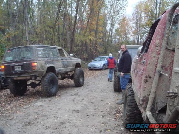 Muddy lifted Jeep Cherokee from rear, with other off-road vehicles and people