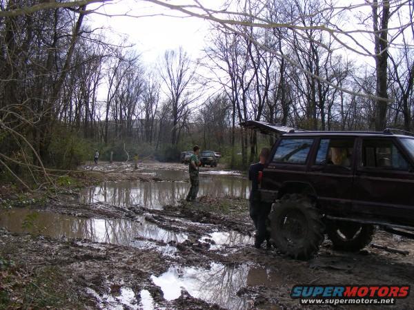 Several off-road vehicles stuck in mud and puddles in a wooded area. One maroon SUV with large tires is prominent