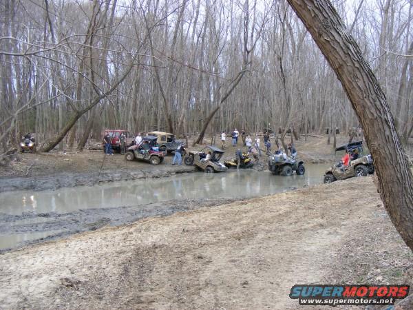 Multiple off-road vehicles navigating a muddy creek surrounded by trees