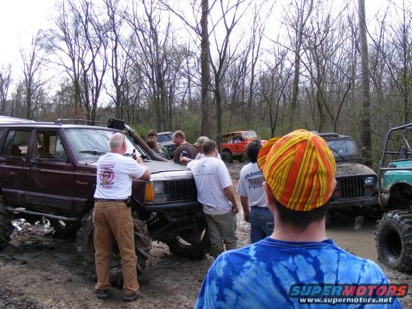 Several off-road vehicles, including a maroon Jeep Cherokee and a black Jeep Wrangler, are in a muddy area with trees in the background. People are gathered around