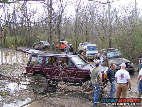 Several off-road vehicles stuck in mud and trees. A maroon Jeep Cherokee is in the foreground