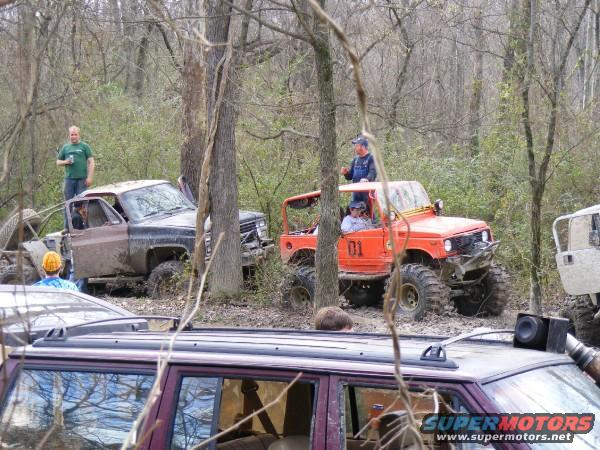 Orange off-road vehicle with number 01 on door, muddy truck, and other vehicles in woods