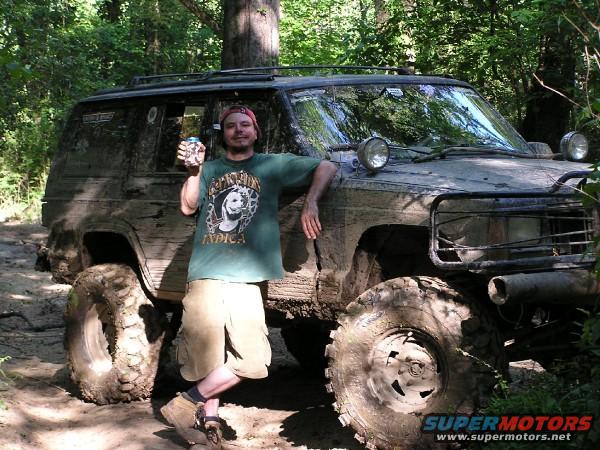 Man with can next to a very muddy SUV with large tires in a wooded area