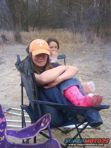 Young woman in orange cap and jeans sits with arms crossed, pink cast on her leg. Another girl sits behind her