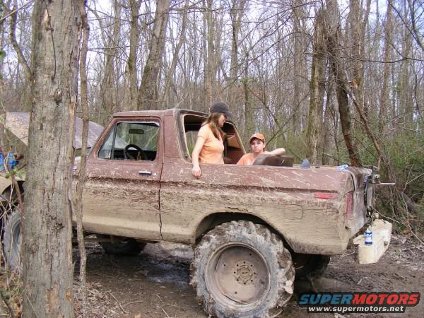 Brown 1978 Ford Bronco covered in mud, with two people inside, parked in woods