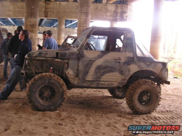 Muddy camouflage SUV parked on sand under a bridge