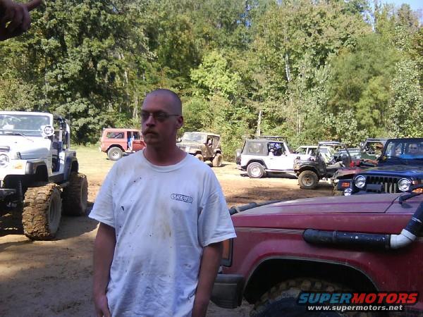 Man in white t-shirt with glasses, standing in a dirt lot with several off-road vehicles and trees in the background. A Supermotors logo is visible on a red vehicle in the foreground