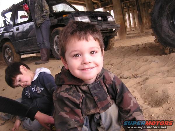 Two young boys in camouflage jackets on sandy ground with a black SUV behind them