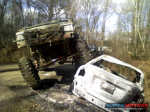 1978 Ford Bronco modified for off-roading, tilted on its side next to a burnt-out SUV