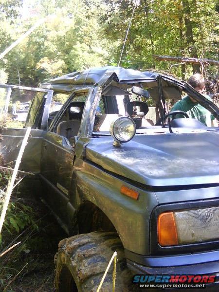 Damaged SUV with dented roof and open doors, stuck in wooded area. Large off-road tires are visible