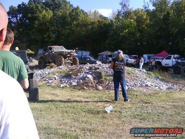 Several off-road vehicles are parked on a rocky incline with trees in the background. People are standing around the vehicles