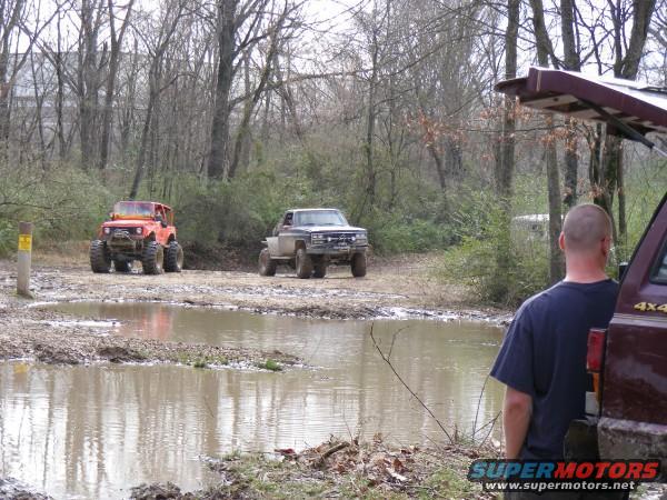 Two off-road vehicles are stuck in mud near trees. A person stands next to a maroon vehicle in the foreground