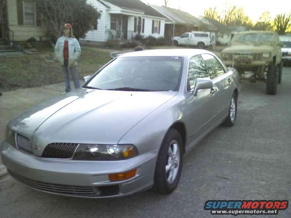 Silver sedan parked on asphalt, front quarter view. A person stands behind it. A lifted off-road vehicle is in the background