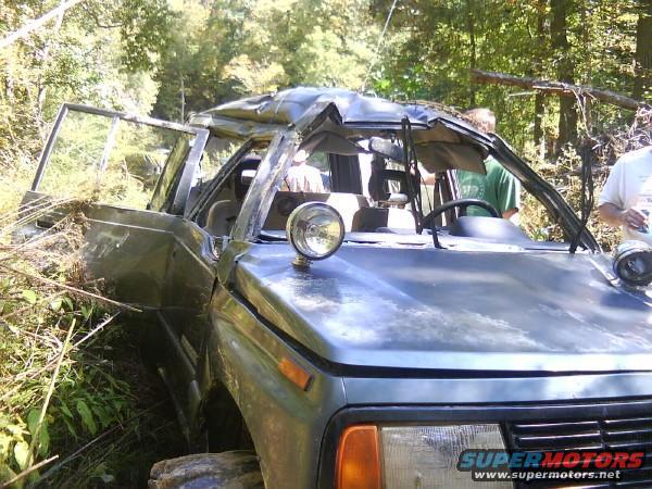 Damaged gray off-road vehicle with open doors and crumpled roof, partially obscured by foliage